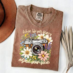Puede incluir: Camiseta marrón con un diseño floral de cámara. El diseño incluye una cámara vintage rodeada de flores coloridas y el texto "When life gets blurry adjust your focus". La camiseta tiene cuello redondo y mangas cortas.