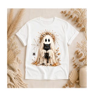 May include: White t-shirt featuring a Halloween-themed design. The graphic depicts a ghost holding a black cat, surrounded by a floral wreath, spider web, and a spider. The design is in shades of beige, brown, and black.