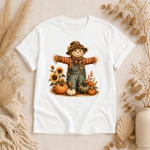 Puede incluir: Una camiseta blanca de manga corta con un gr&aacute;fico central de un espantap&aacute;jaros con sombrero marr&oacute;n, camisa de cuadros y peto vaquero. El espantap&aacute;jaros est&aacute; rodeado de girasoles amarillos, calabazas naranjas, una calabaza blanca y hojas de oto&ntilde;o.