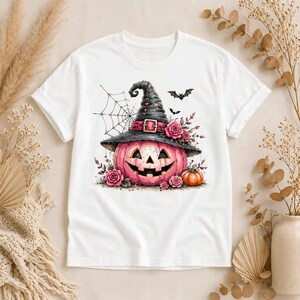 May include: A white short-sleeve t-shirt featuring a graphic of a pink jack-o'-lantern wearing a black witch hat adorned with pink roses. The design includes a spider web, two small black bats, and a smaller orange pumpkin at the base.