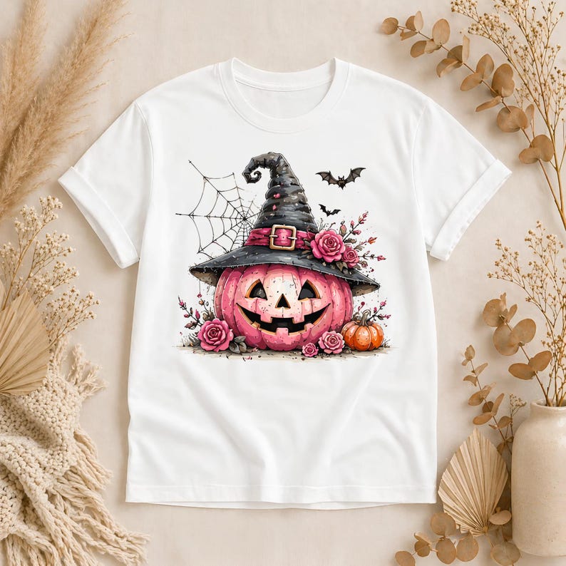 May include: A white short-sleeve t-shirt featuring a graphic of a pink jack-o'-lantern wearing a black witch hat adorned with pink roses. The design includes a spider web, two small black bats, and a smaller orange pumpkin at the base.