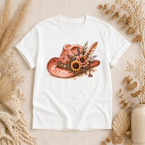 May include: A white short-sleeved t-shirt featuring a graphic print of a pink cowboy hat adorned with a cluster of sunflowers, wildflowers, and a single feather. The hat includes a decorative band and a small dangling charm detail.
