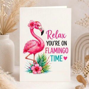 May include: A greeting card featuring a pink flamingo with a flower crown, standing on one leg amidst tropical foliage. The card reads "Relax you're on Flamingo Time" in pink and teal text, with a pink heart.