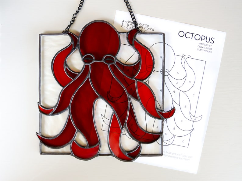 May include: A red stained glass octopus hanging from a chain. The octopus has eight tentacles and is framed in black metal. A pattern for the stained glass is visible below the octopus.