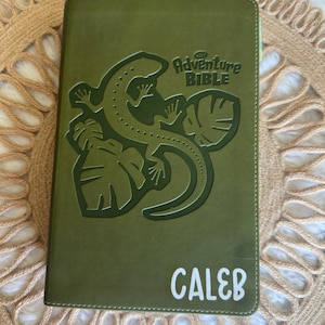 May include: Green leather-bound journal with a lizard and leaves embossed on the cover. The text "The Adventure Bible" is printed in white on the cover. The name "Caleb" is printed in white at the bottom of the cover.