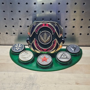 Könnte beinhalten: Ein grüner und goldener Power Rangers Dino Fury Morpher mit einem roten Stern und einer grün-goldenen Basis. Der Morpher ist von sechs kleineren, runden, goldenen und silbernen Münzen mit verschiedenen Symbolen umgeben.