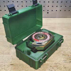 Puede incluir: Una caja de pl&aacute;stico verde abierta, que revela un objeto circular met&aacute;lico con detalles rojos y dorados. El objeto tiene el texto "ZYURANGER" y "KYORYU". La caja est&aacute; sobre una superficie de madera.