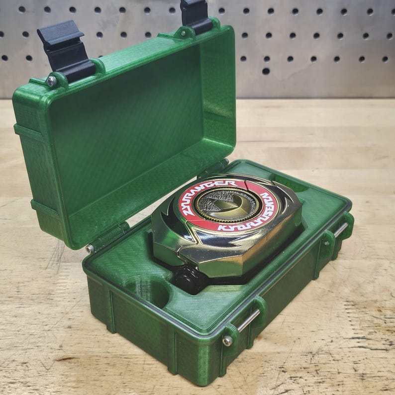 Puede incluir: Una caja de pl&aacute;stico verde abierta, que revela un objeto circular met&aacute;lico con detalles rojos y dorados. El objeto tiene el texto "ZYURANGER" y "KYORYU". La caja est&aacute; sobre una superficie de madera.