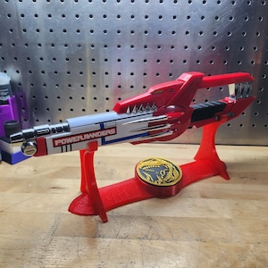 May include: Red and white Power Rangers toy weapon with a silver blade and a red stand. The stand has a gold Power Rangers logo with a dinosaur design.