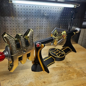Könnte beinhalten: Dekoratives Schwert in Gold und Schwarz mit einem roten Edelstein und einem schwarzen Griff. Das Schwert steht auf einem schwarzen Ständer mit einem goldenen Emblem. Das Design ist detailliert, und das Schwert scheint aus Metall zu sein.