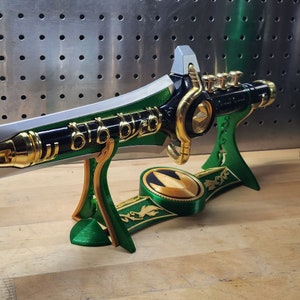 May include: A green and gold Power Rangers sword with a black handle and a green and gold stand. The sword has a gold dragon head on the handle and a gold Power Rangers symbol on the stand.