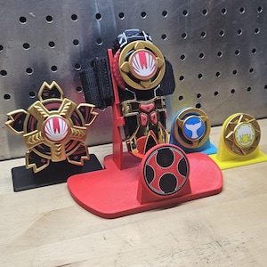 Könnte beinhalten: Eine Sammlung farbenfroher Spielzeug-Accessoires, darunter ein schwarz-goldenes Handgelenkgerät mit roter Basis und mehrere kreisförmige Embleme in Gold, Rot, Blau und Gelb. Die Embleme zeigen verschiedene Symbole.