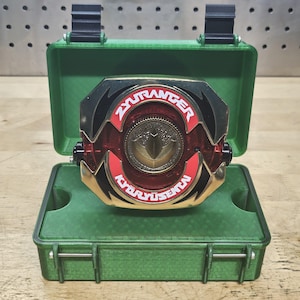 May include: A red, black, and gold ZyuRanger Kyoryu Sentai morpher prop, displayed open inside a green case. The morpher features a gold center emblem and the text "ZyuRanger Kyoryu Sentai". The case has black hinges and latches.