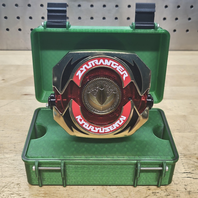 Puede incluir: Un accesorio de morpher ZyuRanger Kyoryu Sentai rojo, negro y dorado, exhibido abierto dentro de un estuche verde. El morpher presenta un emblema central dorado y el texto "ZyuRanger Kyoryu Sentai". La caja tiene bisagras y cierres negros.