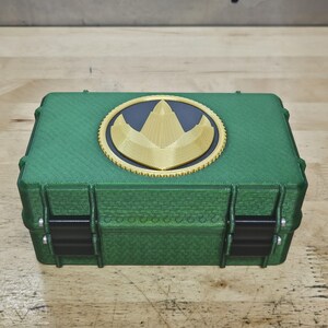 Puede incluir: Caja rectangular verde con un emblema dorado y negro en la tapa. La caja tiene bisagras y cierres negros. El emblema es un dise&ntilde;o dorado sobre fondo negro, dentro de un c&iacute;rculo dorado.
