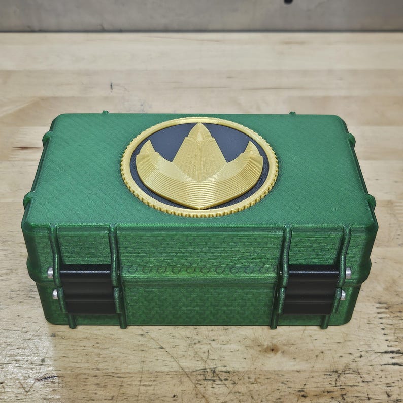 Puede incluir: Caja rectangular verde con un emblema dorado y negro en la tapa. La caja tiene bisagras y cierres negros. El emblema es un dise&ntilde;o dorado sobre fondo negro, dentro de un c&iacute;rculo dorado.