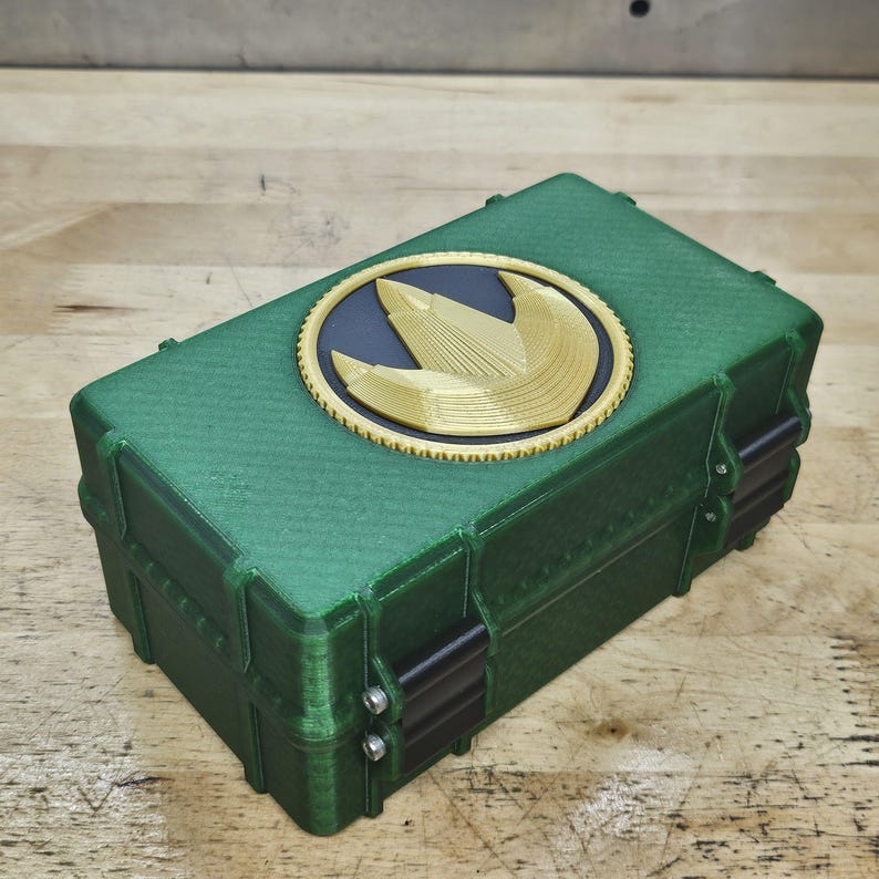 Puede incluir: Una caja rectangular verde con una superficie texturizada y detalles negros. Un emblema dorado y negro est&aacute; centrado en la tapa. La caja tiene un dise&ntilde;o con bisagras y probablemente se utiliza para el almacenamiento.