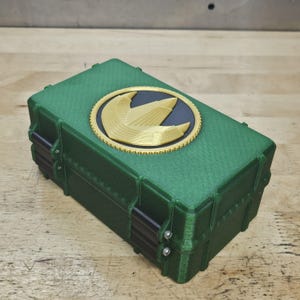Puede incluir: Caja rectangular verde con un emblema dorado y negro en la tapa. El emblema presenta una huella de dinosaurio estilizada. La caja tiene bisagras negras y tornillos plateados. La caja est&aacute; sobre una superficie de madera.