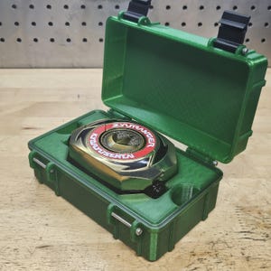 Puede incluir: Un estuche de pl&aacute;stico verde con un dispositivo Power Ranger dorado y rojo en su interior. El dispositivo tiene el texto "ZYURANGER" y "KYORYUSENTA". El estuche est&aacute; abierto, revelando el dispositivo.
