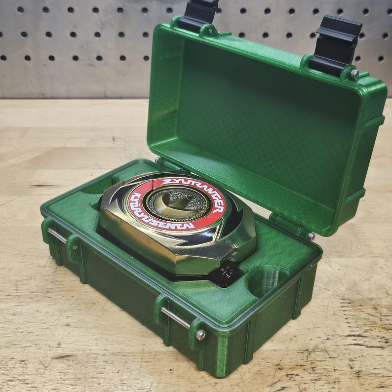 Puede incluir: Un estuche de pl&aacute;stico verde con un dispositivo Power Ranger dorado y rojo en su interior. El dispositivo tiene el texto "ZYURANGER" y "KYORYUSENTA". El estuche est&aacute; abierto, revelando el dispositivo.