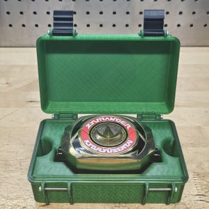 Puede incluir: Un dispositivo morpher ZyuRanger Kyoryu Sentai dorado y negro, con detalles en rojo, descansa dentro de un estuche verde abierto. El estuche tiene una tapa con bisagras y cierres seguros. El texto "ZyuRanger Kyoryu Sentai" es visible.