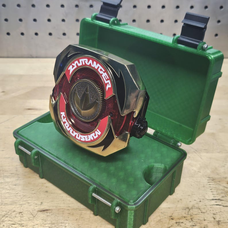 Puede incluir: Un accesorio dorado, rojo y negro con el texto "ZYURANGER" y "KYORYUSENTA" en letras blancas. El accesorio est&aacute; dentro de un estuche verde abierto con cierres negros.