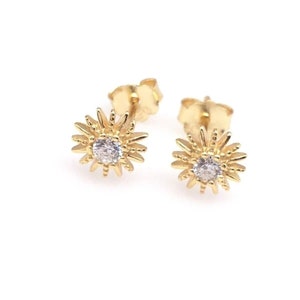 Peut inclure: Une paire de boucles d'oreilles &agrave; clous dor&eacute;es. Chaque boucle d'oreille pr&eacute;sente un motif de soleil avec une pierre pr&eacute;cieuse ronde et transparente au centre. Les boucles d'oreilles sont pos&eacute;es sur un fond blanc, soulignant leurs d&eacute;tails complexes.