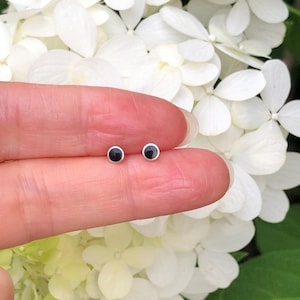 May include: A pair of small, round black stud earrings with silver settings. The earrings are shown on a finger against a background of white flowers.