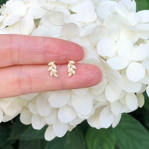 May include: A pair of gold leaf earrings with small clear stones. The earrings are shown on a finger against a background of white flowers.