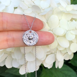 May include: A silver necklace with a round pendant featuring a textured design. The pendant has a small, raised circle in the center.