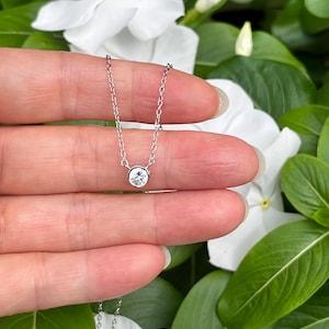 Peut inclure: Un collier délicat de couleur argentée doté d'une pierre ronde, transparente et facettée, montée dans un serti clos lisse. Le pendentif est fixé à une fine chaîne à maillons qui pend verticalement, présentant un design épuré et simple.