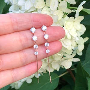 May include: Dangling earrings with clear, round gemstones suspended from delicate rose gold chains. The earrings are displayed against a backdrop of white flowers and green foliage, creating a soft, natural setting. The earrings are a classic jewelry item.