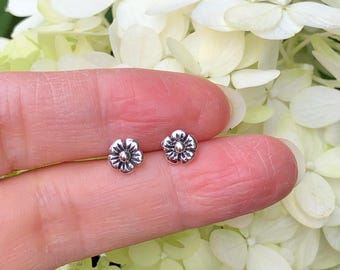 May include: A pair of small, silver-colored flower stud earrings. Each earring features a detailed floral design with multiple petals and a central, textured element. The earrings are displayed against a backdrop of white flowers.