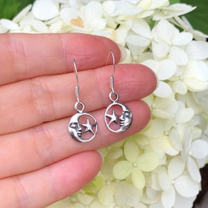 May include: A pair of silver-colored dangle earrings. Each earring features a crescent moon with a face and a star within a circular frame. The earrings are held in a hand against a backdrop of white flowers.