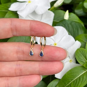 May include: A pair of gold hoop earrings, each featuring a small, dangling teardrop-shaped blue gemstone charm. The earrings are polished and metallic, with a simple hinged closure mechanism visible on the circular hoops.