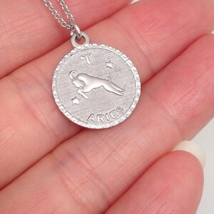 May include: Silver necklace with a round pendant featuring the zodiac symbol for Aries. The pendant has a detailed design with a ram and stars. The pendant also has the word "Aries" inscribed on it.