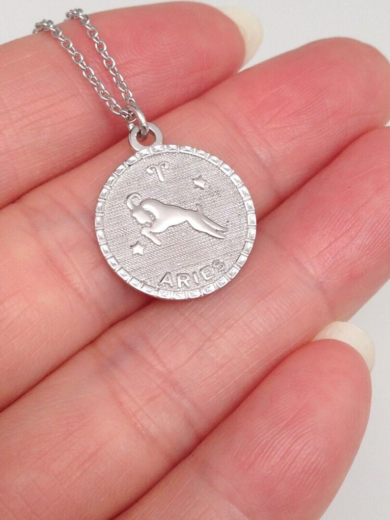 May include: Silver necklace with a round pendant featuring the zodiac symbol for Aries. The pendant has a detailed design with a ram and stars. The pendant also has the word "Aries" inscribed on it.