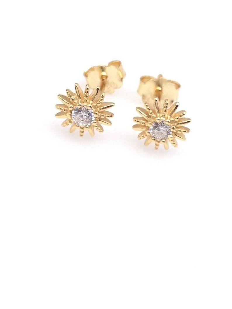Peut inclure: Une paire de boucles d'oreilles &agrave; clous dor&eacute;es. Chaque boucle d'oreille pr&eacute;sente un motif de soleil avec une pierre pr&eacute;cieuse ronde et transparente au centre. Les boucles d'oreilles sont pos&eacute;es sur un fond blanc, soulignant leurs d&eacute;tails complexes.