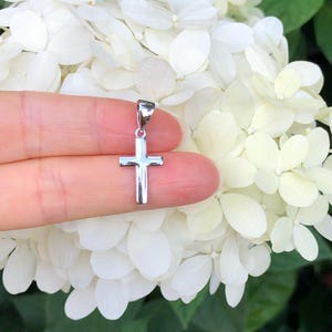 May include: A silver cross pendant charm, held by a finger against a background of white flowers.
