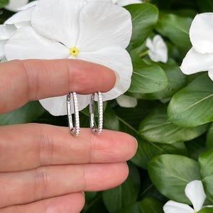May include: A pair of silver-toned hoop earrings featuring a rectangular shape with rounded corners. Each earring is embellished with a row of small, clear, sparkling stones set along the front-facing edge of the metal frame.