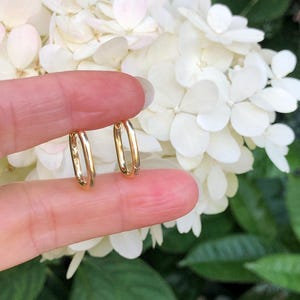 May include: A pair of gold hoop earrings, shown held in a hand against a background of white flowers.