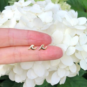 May include: A pair of gold wave earrings. The earrings are small and delicate, and they are shown on a finger against a background of white flowers.