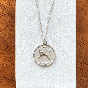 May include: A silver necklace with a circular pendant featuring the zodiac symbol for Aries. The pendant has a detailed design of a ram with the word "Aries" inscribed below.