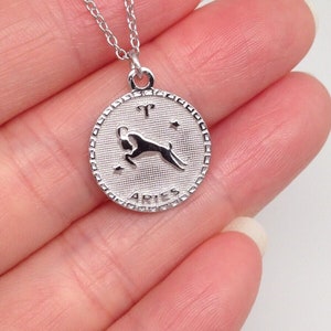 May include: Silver chain necklace with a round pendant featuring the zodiac symbol for Aries. The pendant has a textured surface and the word "Aries" is inscribed around the edge.