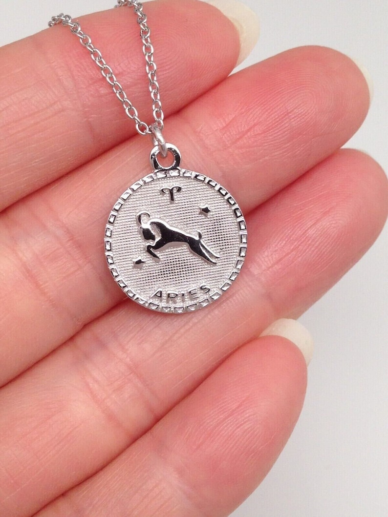 May include: Silver chain necklace with a round pendant featuring the zodiac symbol for Aries. The pendant has a textured surface and the word "Aries" is inscribed around the edge.