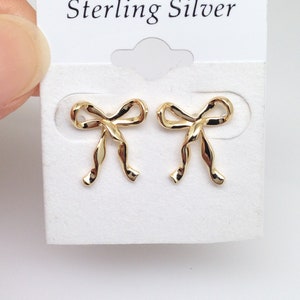 Puede incluir: Un par de pendientes de lazo de plata de ley de color dorado. Los pendientes est&aacute;n en una tarjeta blanca con el texto "Sterling Silver" impreso en ella.