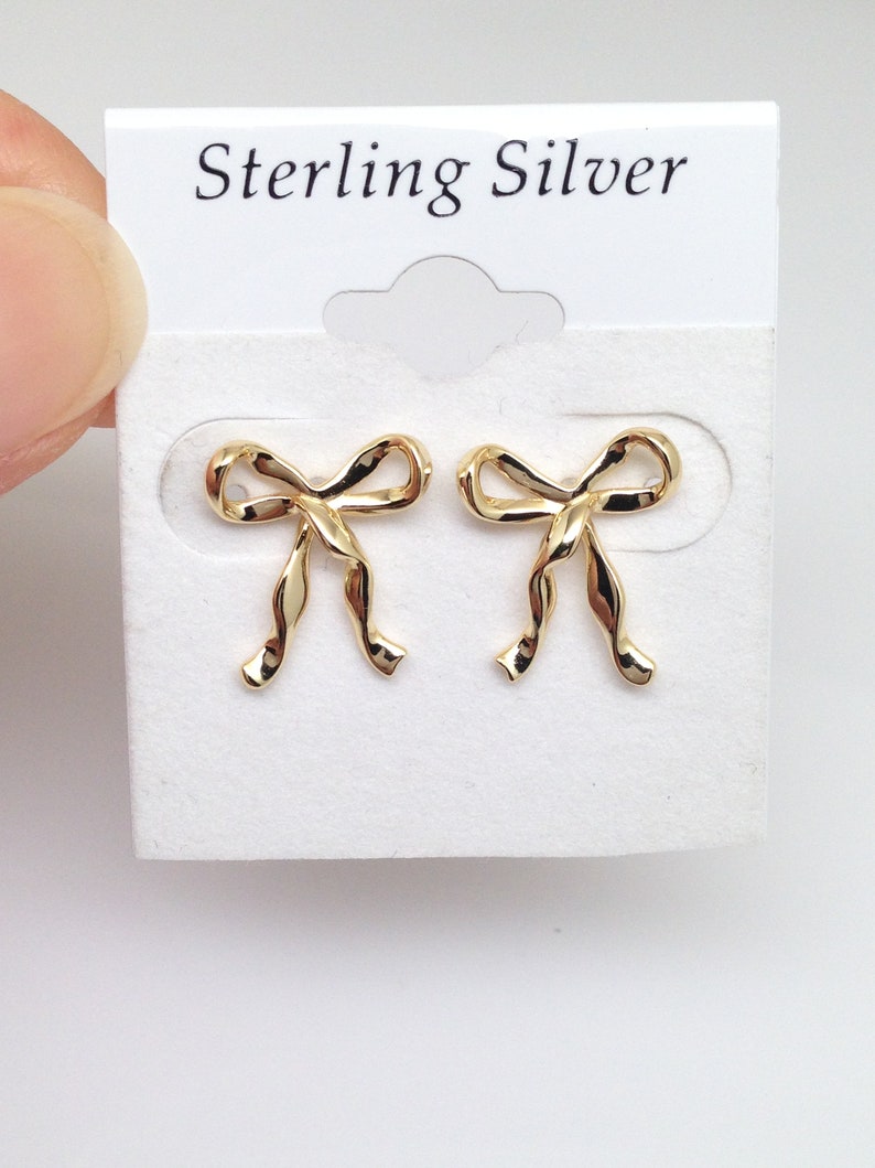 Puede incluir: Un par de pendientes de lazo de plata de ley de color dorado. Los pendientes est&aacute;n en una tarjeta blanca con el texto "Sterling Silver" impreso en ella.