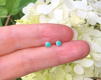 May include: A pair of small, round turquoise earrings with silver posts. The earrings are shown on a person's finger, with a white flower in the background.