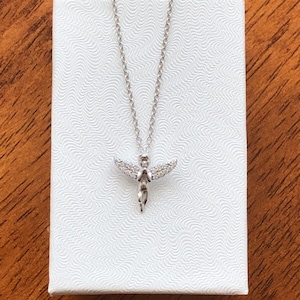 Puede incluir: Un collar de cadena de plata con un peque&ntilde;o colgante de &aacute;ngel brillante. El &aacute;ngel tiene alas y lleva un vestido. El collar est&aacute; en una caja blanca.