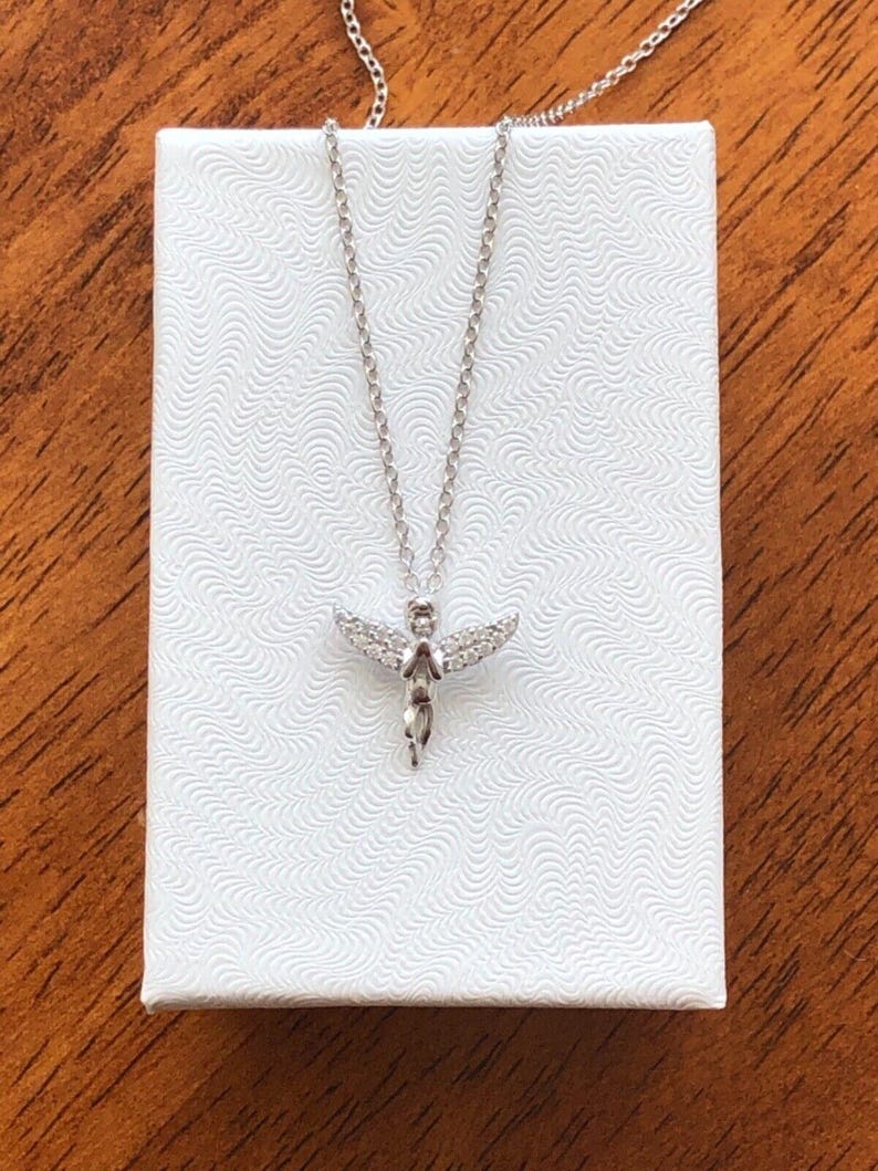 Puede incluir: Un collar de cadena de plata con un peque&ntilde;o colgante de &aacute;ngel brillante. El &aacute;ngel tiene alas y lleva un vestido. El collar est&aacute; en una caja blanca.
